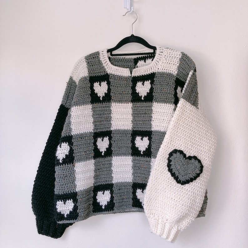 Pu&ograve; includere: Un maglione a uncinetto grigio e bianco con accenti a forma di cuore neri. Il maglione ha un motivo a scacchi e una vestibilit&agrave; ampia.