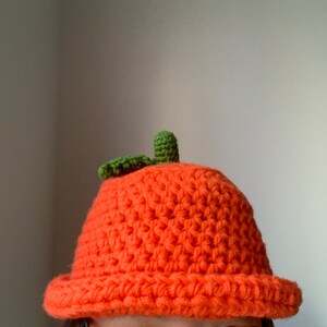 Pu&ograve; includere: Un cappello arancione all'uncinetto a forma di zucca con un gambo verde.