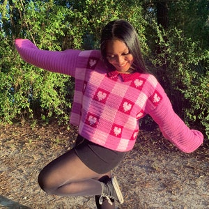Pu&ograve; includere: Un maglione di uncinetto rosa e bianco con un motivo a forma di cuore. Il maglione ha un colletto e maniche lunghe. La modella indossa collant neri e scarpe da ginnastica nere.