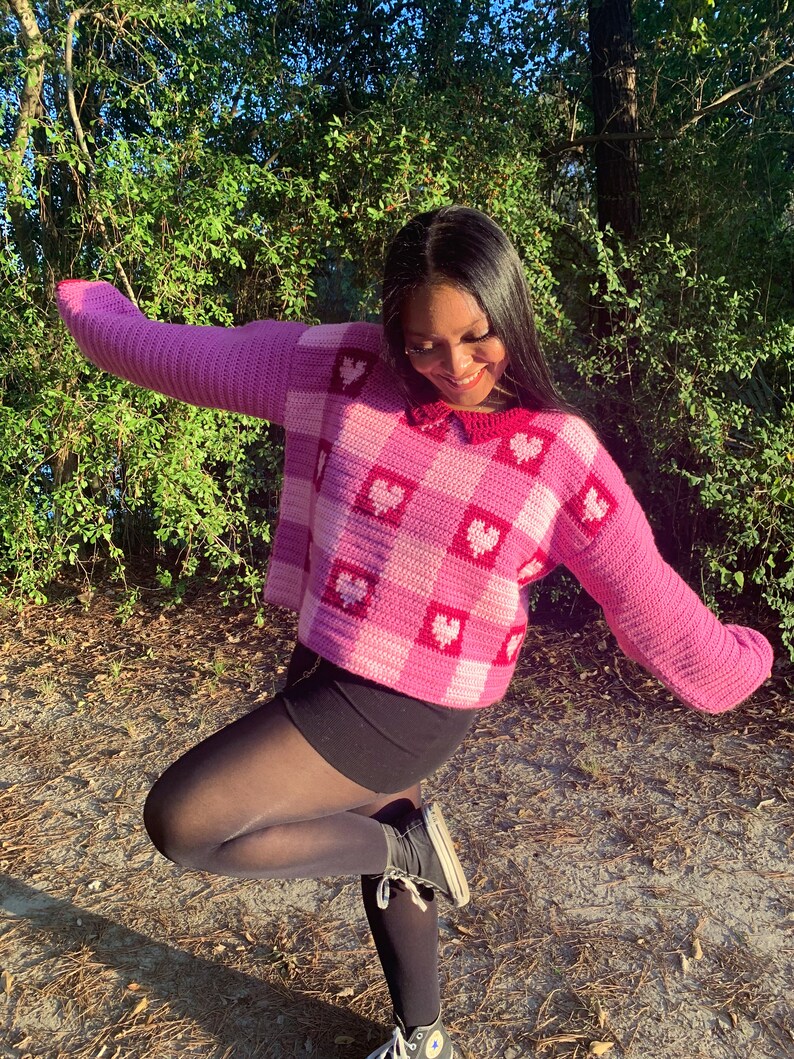 Pu&ograve; includere: Un maglione di uncinetto rosa e bianco con un motivo a forma di cuore. Il maglione ha un colletto e maniche lunghe. La modella indossa collant neri e scarpe da ginnastica nere.