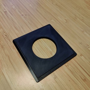 May include: A black plastic square with a round hole in the center. The square is on a light brown wooden surface.
