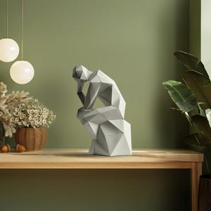 Puede incluir: Una escultura geométrica gris de una persona en posición sentada y pensativa. La escultura está sobre una mesa de madera, con una pared verde de fondo. La escultura mide aproximadamente 30 cm de altura.