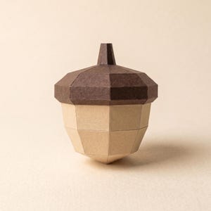 Puede incluir: Una escultura geométrica de bellota hecha de papel con una tapa de color marrón oscuro y un cuerpo de color tostado claro. El objeto está construido con múltiples paneles de papel planos y angulares que crean una forma tridimensional facetada.