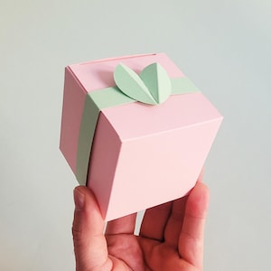 May include: A pink gift box with a green paper bow and a heart-shaped cutout. The box is being held in a hand.