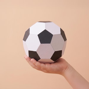 May include: A geometric paper soccer ball featuring a classic pattern of alternating black pentagons and white hexagons. The structure is constructed from flat, angular panels, creating a faceted, three-dimensional sphere with clean, sharp edges.