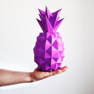 May include: A purple origami pineapple, made of paper, is held in a person's hand. The pineapple is a geometric shape with many facets.