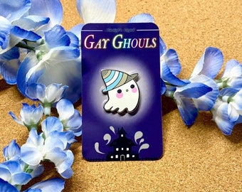 May include: A blue and white striped ghost wearing a witch's hat with a pink face and a white body. The ghost is on a purple card with the text "Gay Ghouls" in white letters. The card also has a black silhouette of a house with white windows.