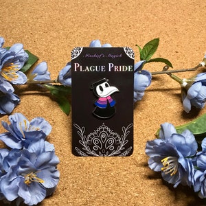 May include: A black enamel pin depicting a plague doctor with a white beak and hat. The doctor's attire incorporates the colours of the bisexual pride flag. The pin is displayed on a black card with the text "PLAGUE PRIDE".