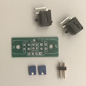 May include: An assortment of electronic components, including a green circuit board with text, two black connectors with silver pins, two blue connectors, and a black and silver connector. The components are arranged on a light gray surface.