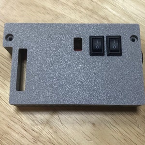 Op de afbeelding: Een grijze kunststof behuizing met twee zwarte schakelaars en een rechthoekige uitsparing. De behuizing heeft een gestructureerd oppervlak.