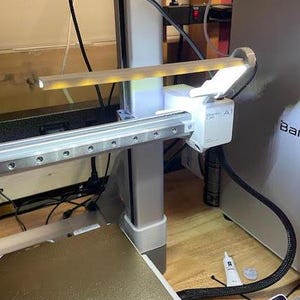May include: A close-up of a 3D printer with a white light attached to the arm. The light is on and illuminating the print bed. The printer is on a wooden surface.