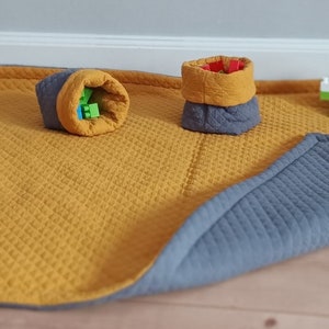 Könnte beinhalten: Eine Wendespielmatte mit gesteppter Textur, senfgelb auf der einen und grau auf der anderen Seite. Zwei Stoffbehälter, einer gerollt und einer aufrecht, enthalten bunte Spielzeugblöcke. Eine kleine Spielzeugblockstruktur liegt auf der Matte.