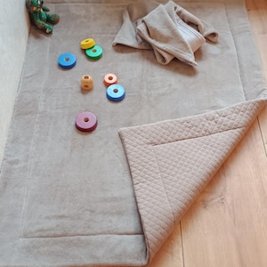 Könnte beinhalten: Eine hellbraune Baby-Spielmatte mit gestepptem Rand und einer gefalteten Ecke. Ein Stapel bunter Holzringe und ein kleiner Teddybär liegen auf der Matte. Eine gefaltete Decke liegt auf der Matte.