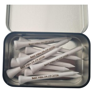 May include: A metal tin filled with white golf tees. Each tee is inscribed with "Best Man 04.07.2026" in a dark font. The tin is open, revealing the contents, and is set against a white background.