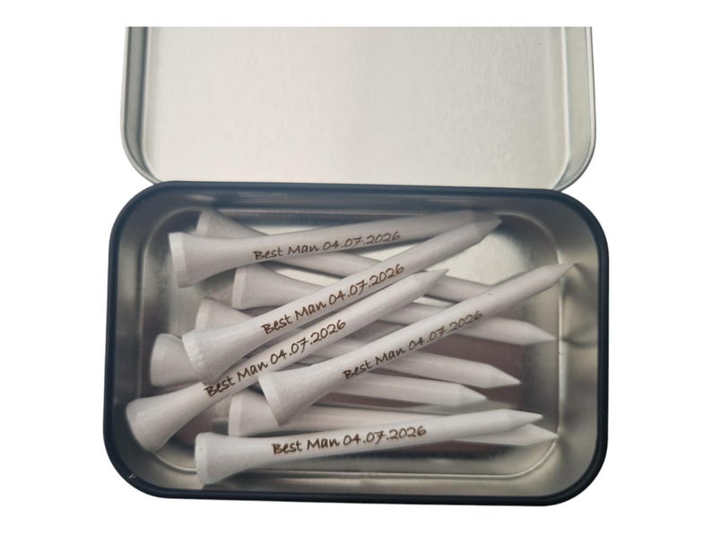 May include: A metal tin filled with white golf tees. Each tee is inscribed with "Best Man 04.07.2026" in a dark font. The tin is open, revealing the contents, and is set against a white background.