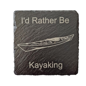 Pode incluir: Uma base de ard&oacute;sia cinza escura quadrada com o texto "I'd Rather Be Kayaking" e um desenho de linha de um caiaque. O texto e o caiaque s&atilde;o gravados em uma cor mais clara, criando um contraste.