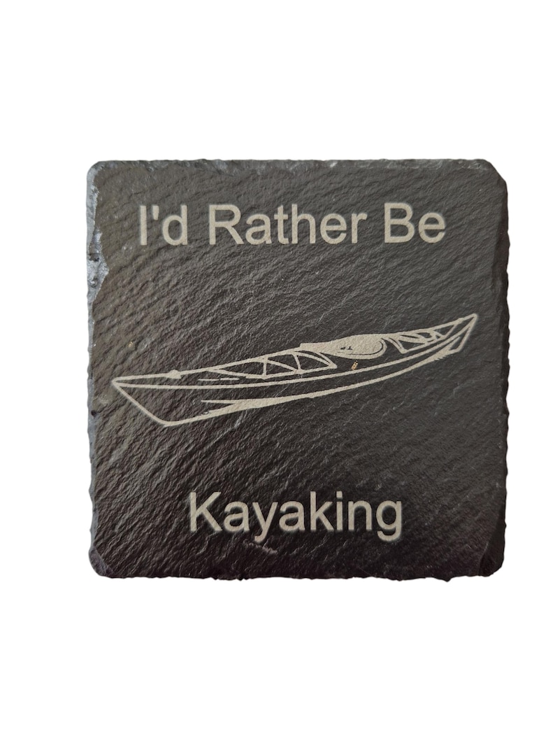 Pode incluir: Uma base de ard&oacute;sia cinza escura quadrada com o texto "I'd Rather Be Kayaking" e um desenho de linha de um caiaque. O texto e o caiaque s&atilde;o gravados em uma cor mais clara, criando um contraste.