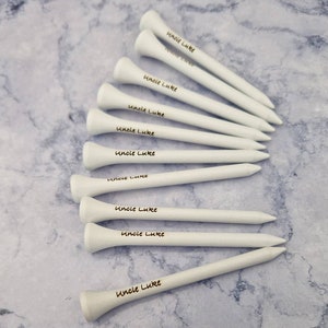 May include: A collection of white wooden golf tees, each with the text "Uncle Luke" printed in black. The tees are arranged on a light gray and white marbled surface.