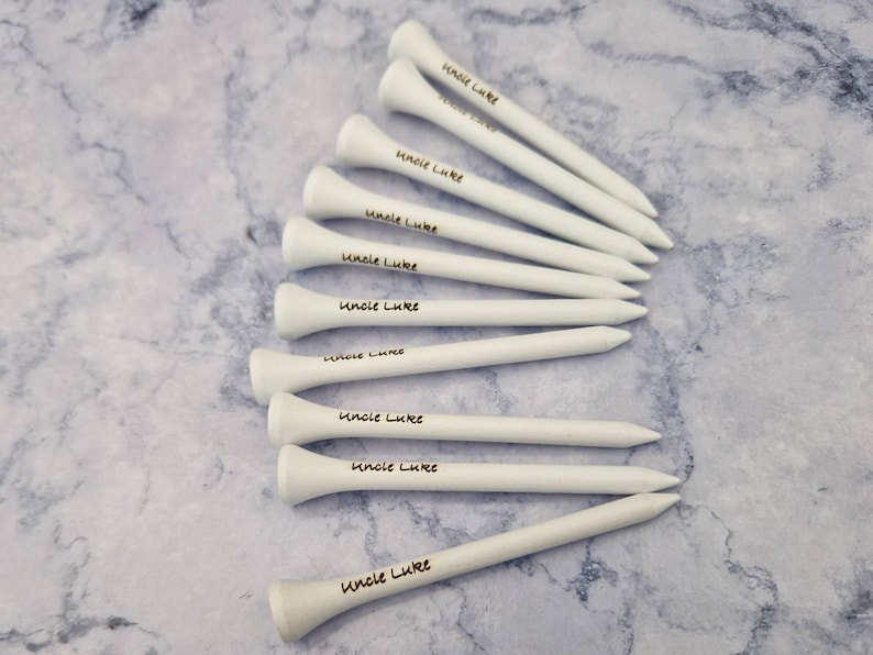 May include: A collection of white wooden golf tees, each with the text "Uncle Luke" printed in black. The tees are arranged on a light gray and white marbled surface.