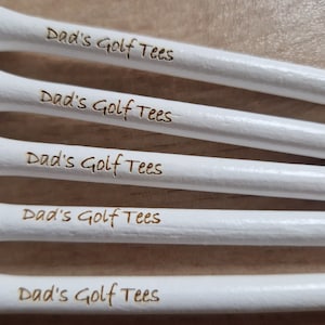 May include: Close-up of five white golf tees with the text "Dad's Golf Tees" etched in a brown color. The tees are arranged in a parallel, slightly angled pattern, suggesting a golf-related theme.