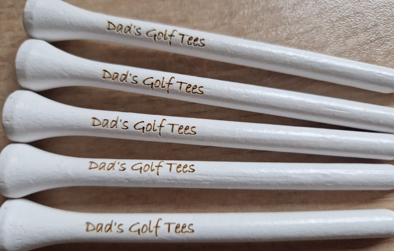 May include: Close-up of five white golf tees with the text "Dad's Golf Tees" etched in a brown color. The tees are arranged in a parallel, slightly angled pattern, suggesting a golf-related theme.