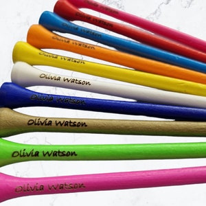May include: Ten personalised golf tees in a variety of colours, including pink, red, orange, yellow, white, blue, green, and brown. Each tee is engraved with the name "Olivia Watson".