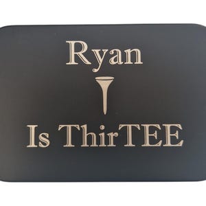 Op de afbeelding: Een zwarte rechthoekige plaat met afgeronde hoeken. De tekst "Ryan Is ThirTEE" is gegraveerd in een lichte kleur, met een golf-tee symbool tussen de namen en het nummer. De plaat is waarschijnlijk een cadeau of een gedenkitem.