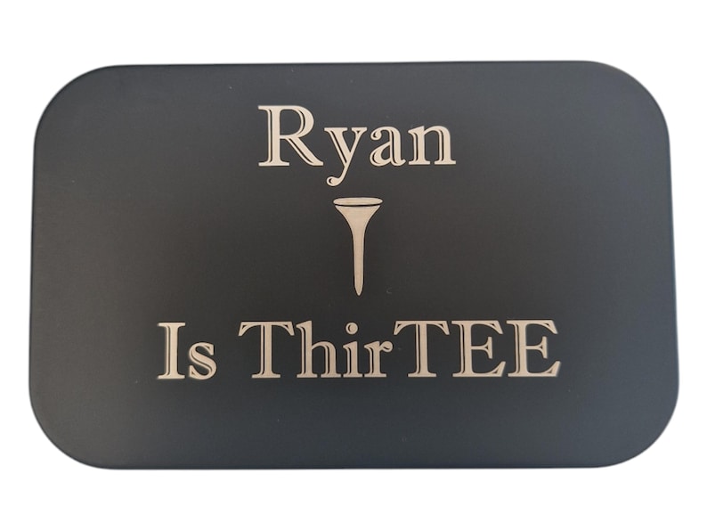 Op de afbeelding: Een zwarte rechthoekige plaat met afgeronde hoeken. De tekst "Ryan Is ThirTEE" is gegraveerd in een lichte kleur, met een golf-tee symbool tussen de namen en het nummer. De plaat is waarschijnlijk een cadeau of een gedenkitem.