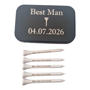 May include: A dark gray rectangular tin with the text "Best Man" and the date "04.07.2026" in silver lettering. Below are five white golf tees, each with the text "Best Man 04.07.2026".