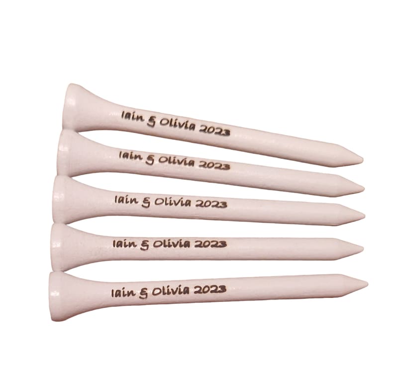 May include: Five white wooden golf tees. Each tee has the text "Iain & Olivia 2023" printed in black. The tees are arranged in a diagonal stack, with the tips pointing to the right.