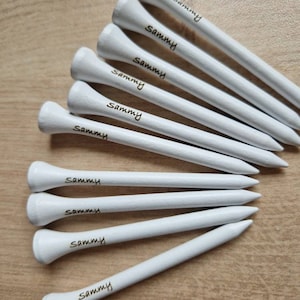 May include: A collection of white golf tees with the name "Sammy" printed in black. The tees are arranged on a light brown wooden surface, showcasing their pointed ends and rounded tops.