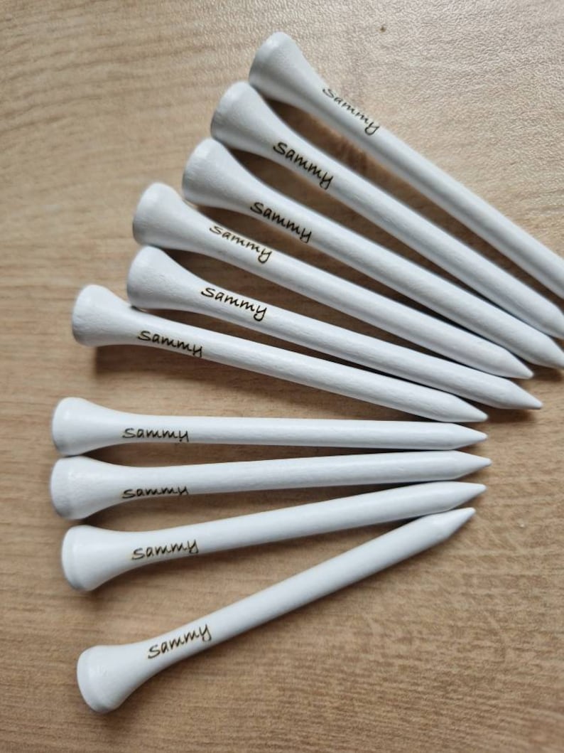 May include: A collection of white golf tees with the name "Sammy" printed in black. The tees are arranged on a light brown wooden surface, showcasing their pointed ends and rounded tops.