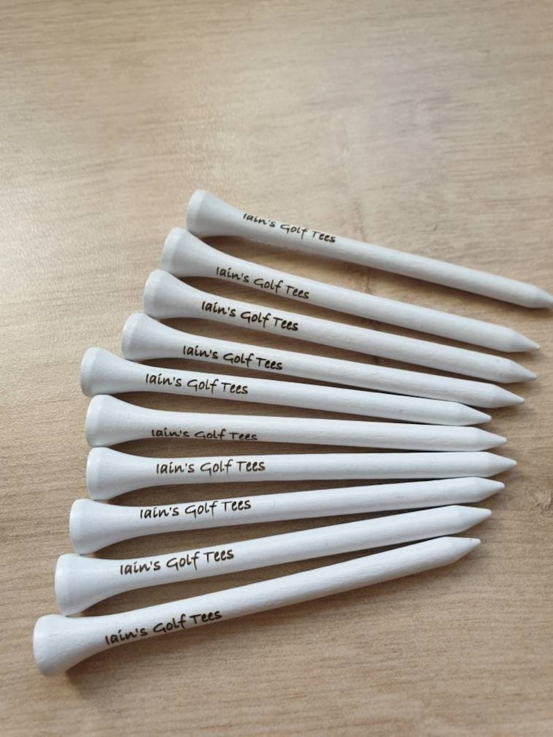 May include: A collection of white golf tees, each with a rounded top and a pointed end. The text "Iain's Golf Tees" is written in black script on each tee. The tees are arranged on a light brown wooden surface.