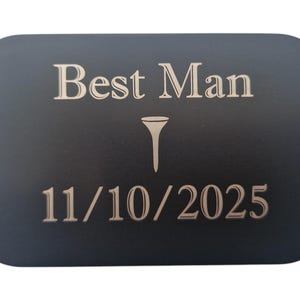 May include: A black rectangular plaque with rounded corners, featuring the words "Best Man" above a golf tee symbol and the date "11/10/2025" in a light beige color. The text is centered on the plaque.