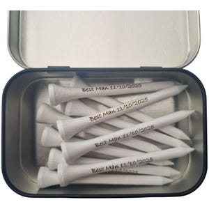 May include: A metal tin filled with white golf tees. Each tee is engraved with the text "Best Man 11/10/2025". The tin is open, revealing the contents. The tees are arranged in a pile.