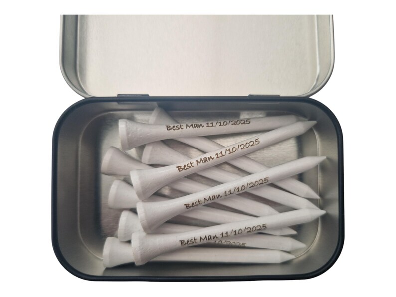 May include: A metal tin filled with white golf tees. Each tee is engraved with the text "Best Man 11/10/2025". The tin is open, revealing the contents. The tees are arranged in a pile.