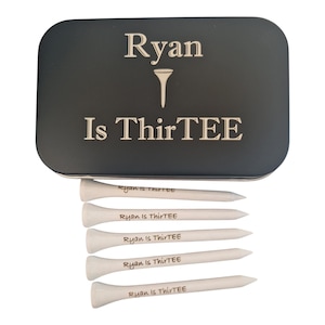 Op de afbeelding: Een zwarte metalen golf tee box met de tekst "Ryan Is ThirTEE" in het wit. Daaronder zes witte golf tees, elk met "Ryan is ThirTEE" erop gedrukt. Een cadeau set voor een golfer.