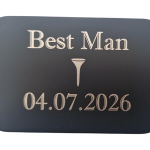 May include: A dark gray rectangular plaque with rounded corners. The text "Best Man" is at the top, with a golf tee symbol below it, and the date "04.07.2026" at the bottom. The text and symbol are in a lighter color.