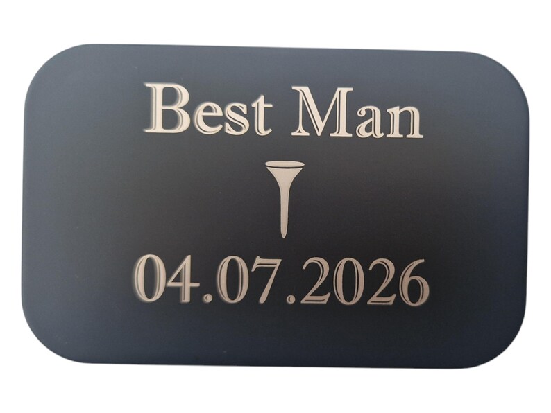 May include: A dark gray rectangular plaque with rounded corners. The text "Best Man" is at the top, with a golf tee symbol below it, and the date "04.07.2026" at the bottom. The text and symbol are in a lighter color.