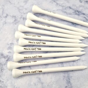 May include: A collection of white golf tees, each with a rounded top and a pointed end. The text "Paul's Golf Tees" is printed on each tee. The tees are arranged on a light blue and white marbled surface.