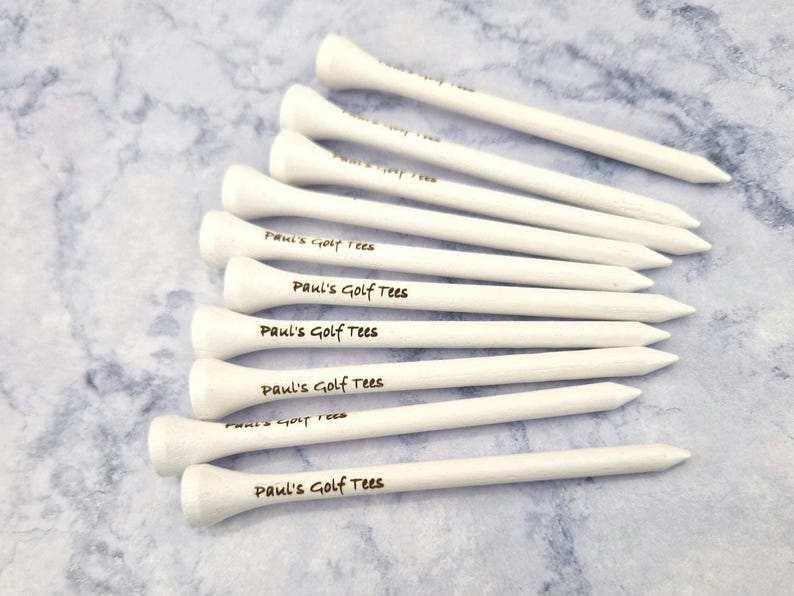 May include: A collection of white golf tees, each with a rounded top and a pointed end. The text "Paul's Golf Tees" is printed on each tee. The tees are arranged on a light blue and white marbled surface.