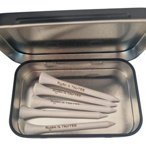 Op de afbeelding: Een open zilveren metalen doos met vijf witte golftees. Elke tee is beschreven met de tekst "Ryan is ThirtEE" in handschrift. De tees zijn in de doos gerangschikt.