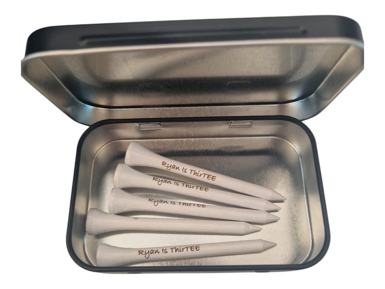 Op de afbeelding: Een open zilveren metalen doos met vijf witte golftees. Elke tee is beschreven met de tekst "Ryan is ThirtEE" in handschrift. De tees zijn in de doos gerangschikt.