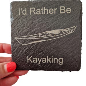 Pode incluir: Uma base para copos quadrada de ard&oacute;sia cinza escuro com o texto "I'd Rather Be Kayaking" e uma ilustra&ccedil;&atilde;o de caiaque gravada em branco. A base para copos &eacute; segurada por uma pessoa com verniz de unhas vermelho.