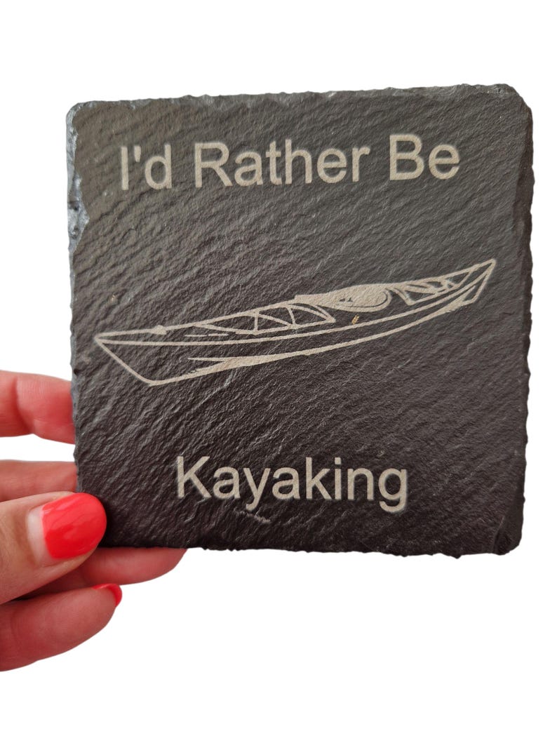 Pode incluir: Uma base para copos quadrada de ard&oacute;sia cinza escuro com o texto "I'd Rather Be Kayaking" e uma ilustra&ccedil;&atilde;o de caiaque gravada em branco. A base para copos &eacute; segurada por uma pessoa com verniz de unhas vermelho.