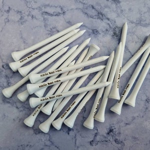 May include: A collection of white golf tees, each with the text "Kevin Best Man" printed in black. The tees are arranged on a light blue and gray marbled surface, showcasing a personalized gift for a golf enthusiast.