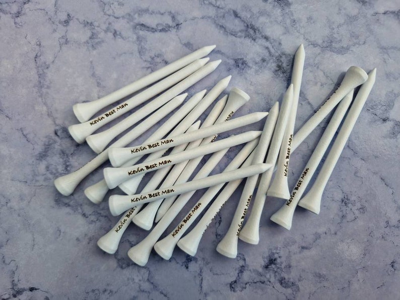 May include: A collection of white golf tees, each with the text "Kevin Best Man" printed in black. The tees are arranged on a light blue and gray marbled surface, showcasing a personalized gift for a golf enthusiast.