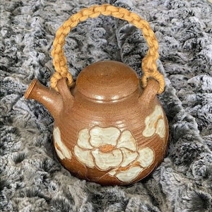 May include: A brown ceramic teapot with a beige rope handle and white floral designs. The teapot is sitting on a gray faux fur surface.