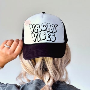 May include: A black and white trucker hat with the text "Vacay Vibes" in a retro font. The hat has a black mesh back and a white front.