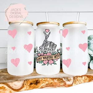 May include: Three frosted glass tumblers with bamboo lids and straws. The center tumbler features a skeleton hand making a peace sign with a heart above it and a floral design with the text "Love Yourself First". The other two tumblers have a pink heart pattern.